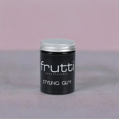 Czarny pojemnik z gumą do stylizacji włosów Frutti Professional Styling Gum, z srebrną nakrętką i białym logo marki, na tle w odcieniach pastelowego różu.